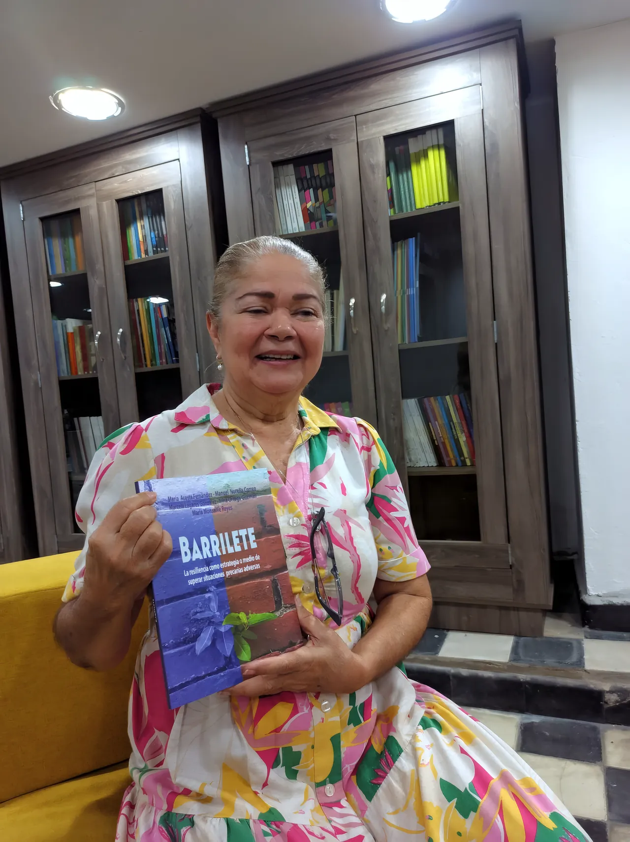 La PhD María del Socorro Acosta, sonriente, sostiene el libro «Barrilete» frente a una biblioteca